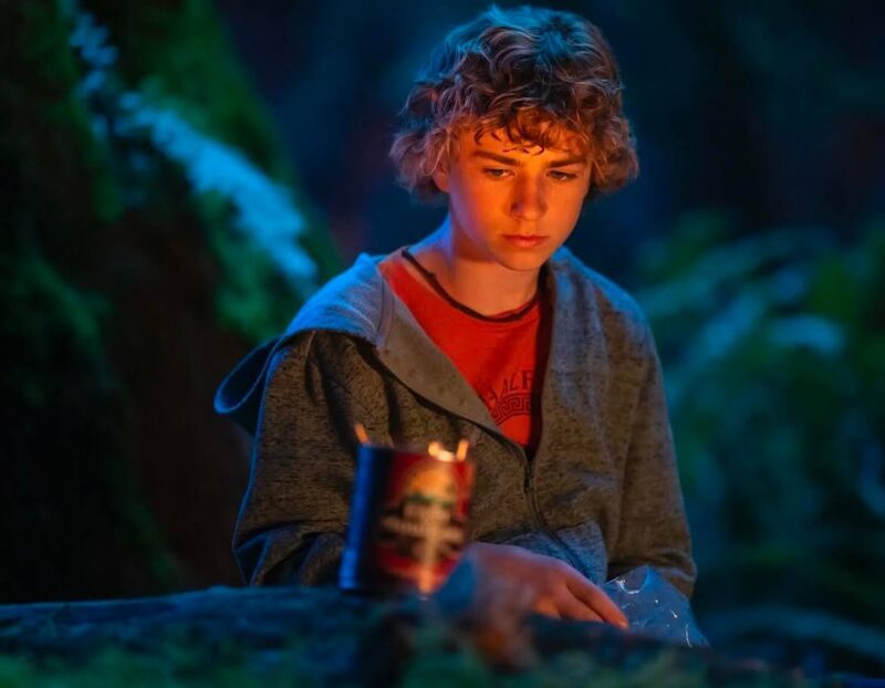 A imagem mostra um menino adolescente sentado à noite em uma floresta. Ele tem cabelos cacheados claros e usa uma camiseta laranja por baixo de um casaco cinza com capuz. O garoto olha para baixo com expressão pensativa, iluminado por uma luz quente que parece vir de uma fogueira fora de cena. À sua frente, sobre um tronco, há uma lata aberta e um saco plástico transparente. O fundo é escuro, com tons azulados e verdes, referindo-se a vegetação, criando um clima silencioso e introspectivo.