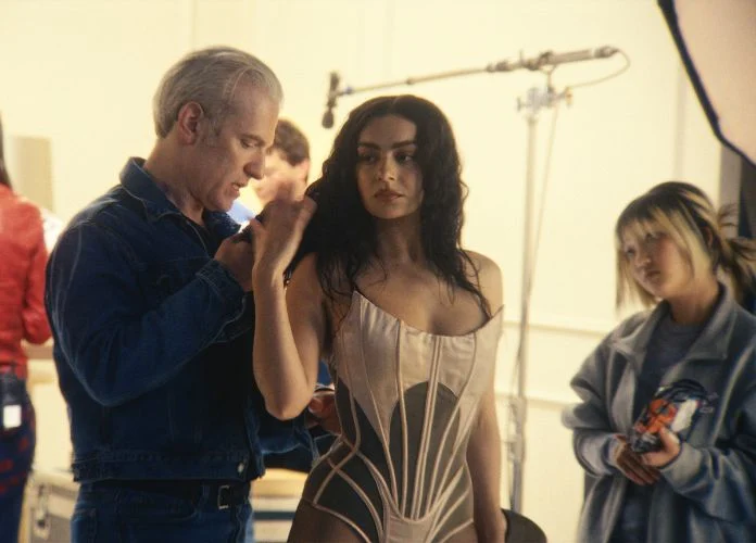Charli XCX, uma mulher branca com cabelo ondulado escuro, está em um estúdio com um corset claro, enquanto um homem loiro e branco, vestido de jeans, está arrumando a sua roupa. Do outro lado, tem uma mulher asiática com um pacote na mão, olhando para eles. No fundo, há outras duas pessoas desfocadas.