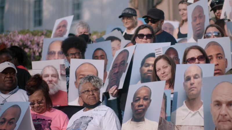 Cena de Alabama: Presos do Sistema. Há um grupo de pessoas reunidas em uma manifestação. Elas seguram grandes retratos de diversos homens com expressões sérias em um fundo neutro. 