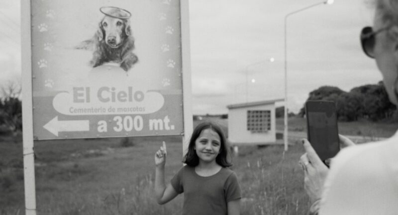 Cena de A Mensageira. Em primeiro plano há as mãos e parte do rosto de Myriam segurando o celular e fotografando a cena. No centro, Anika sorri e aponta para uma placa que exibe a ilustração de um cachorro com auréola e os dizeres "El Cielo - Cementerio de mascotas a 300 mt" com uma seta para a esquerda. 