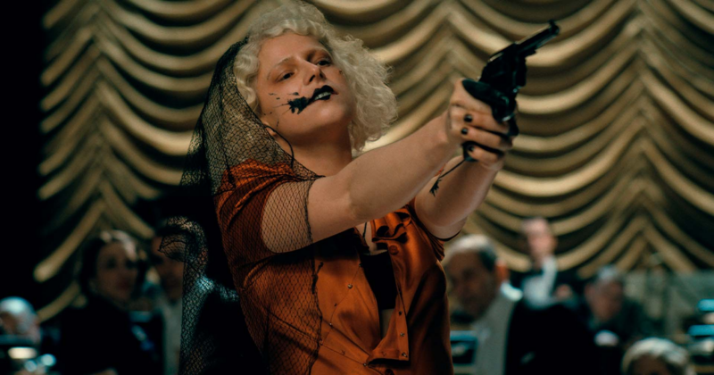 Mulher loira com véu preto e vestido vermelho aponta um revólver em um palco, diante de uma plateia em um ambiente com cortinas douradas