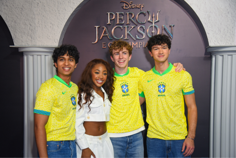 Foto da premiere brasileira da série Percy Jackson e os Olimpianos. Na imagem, quatro jovens atores posam sorridentes em frente a um painel com o logo da série. Dois rapazes com cabelos escuros e curtos vestem uniformes amarelos da seleção brasileira de futebol. Entre eles, uma jovem negra com cabelos longos e ondulados usa uma jaqueta cropped branca e calça branca. Ao lado deles, um rapaz branco de cabelos loiros e encaracolados sorri para a câmera. Todos os rapazes estão com a camisa da seleção brasileira