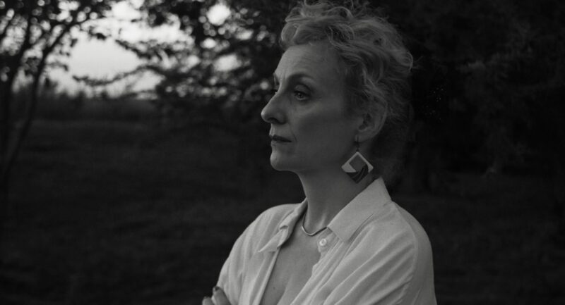 Cena do filme A Mensageira Myriam, uma mulher branca de cabelos loiros cacheados, está de perfil, com os braços cruzados, olhando para a paisagem. Ela usa uma camisa clara aberta no colo e brincos grandes. A expressão dela é séria.