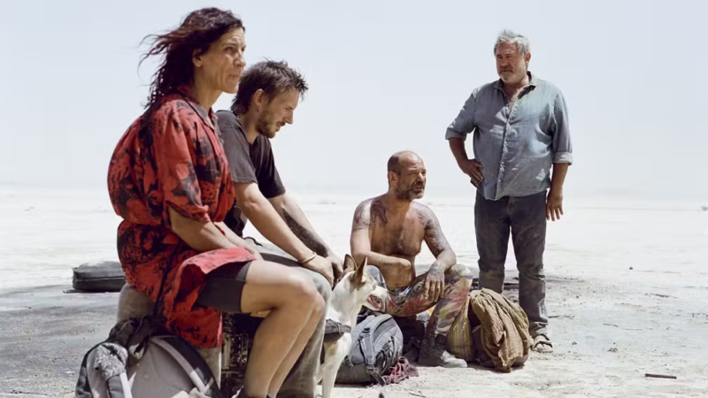 Cena de Sirât. Quatro pessoas e um cachorro estão em uma vasta planície branca e árida, sob uma luz solar intensa. Em primeiro plano, uma mulher de cabelos escuros e vestido estampado vermelho observa o horizonte. Ao lado dela, dois homens estão sentados no chão junto a mochilas e um cachorro branco de pequeno porte. À direita, um homem grisalho de camisa azul está de pé, com a mão na cintura, observando os outros. O clima é de exaustão e desolação.