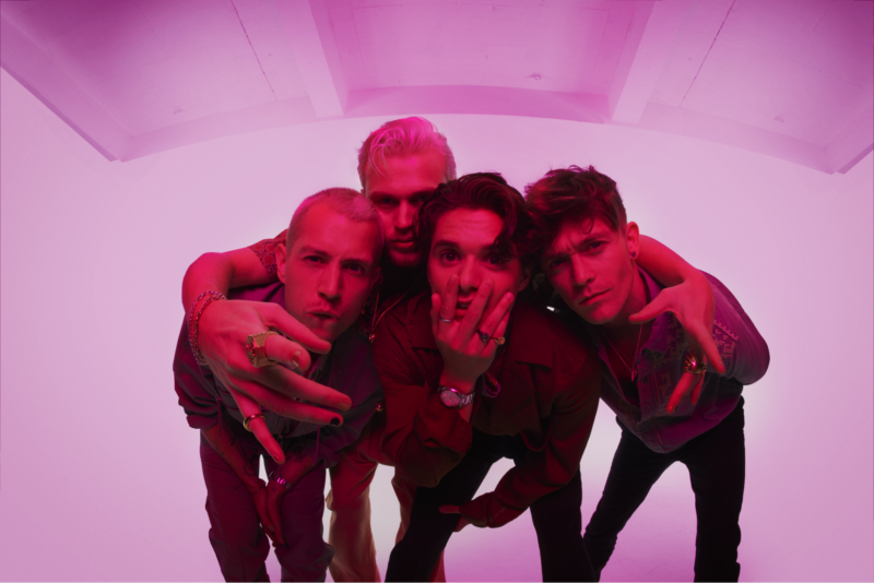 Na fotografia, um grupo de quatro homens caucasianos abraçados e agrupados em um estúdio com iluminação rosa vibrante. Eles vestem roupas casuais, com camisas em tons escuros. Suas expressões faciais variam, com alguns parecendo sérios e outros com leve tom de análise e espanto. A iluminação rosa banha todo o cenário, criando uma atmosfera estilizada e quase surreal. A composição é centralizada, com os rostos dos homens em foco, ocupando a maior parte do quadro. O fundo branco e liso contrasta com as roupas escuras e a luz rosa, concentrando a atenção nos artistas.