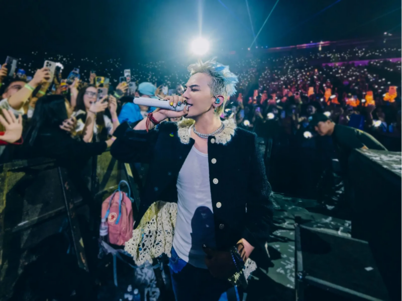 G-DRAGON caminhando em um show em meio a multidão. Fotografia retangular, ao fundo seus fãs no estádio, com o cantor ao centro. G-DRAGON é um homem asiático, de cabelos platinados com detalhes em azul. Ele veste uma regata branca, um sobretudo preto com botões brancos e detalhes de crochê na gola. Ele passa pela multidão enquanto canta e segura um microfone branco à sua frente.