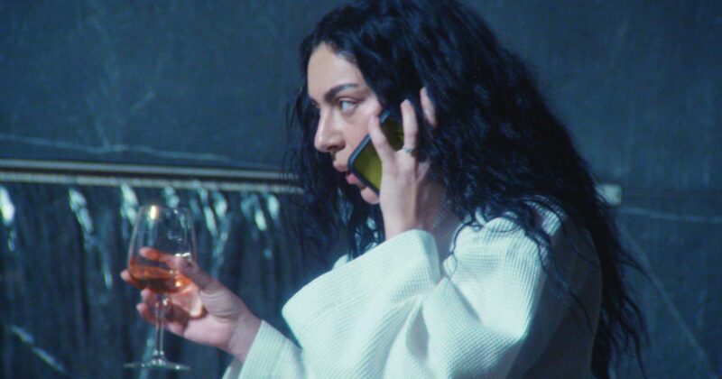 Charli XCX, uma mulher branca com cabelo ondulado escuro, está virada para a esquerda enquanto fala no telefone e na outra mão tem uma taça com bebida. Ela está de roupão branco e o ambiente é escuro.
