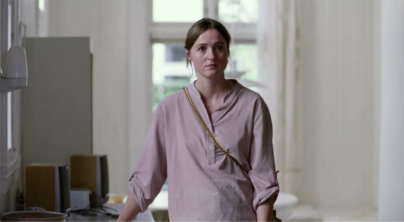 Cena do filme Valor Sentimental. Na imagem, há uma mulher branca de cabelos castanhos em uma casa. Ela veste uma blusa com tons terrosos e uma bolsa dourada utilizada na transversal.