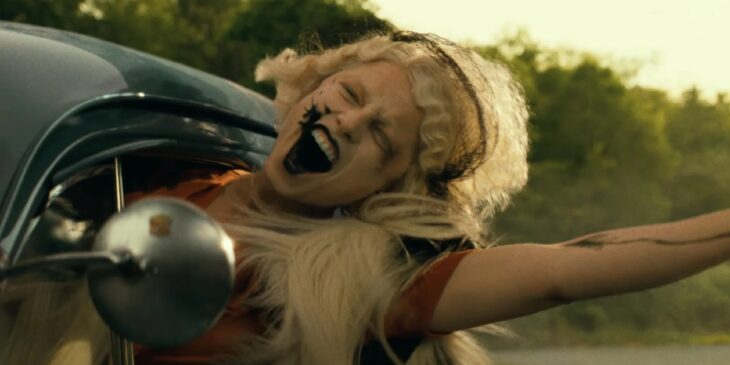  Mulher loira com maquiagem preta borrada grita pela janela de um carro em movimento, com os braços abertos e vento no cabelo