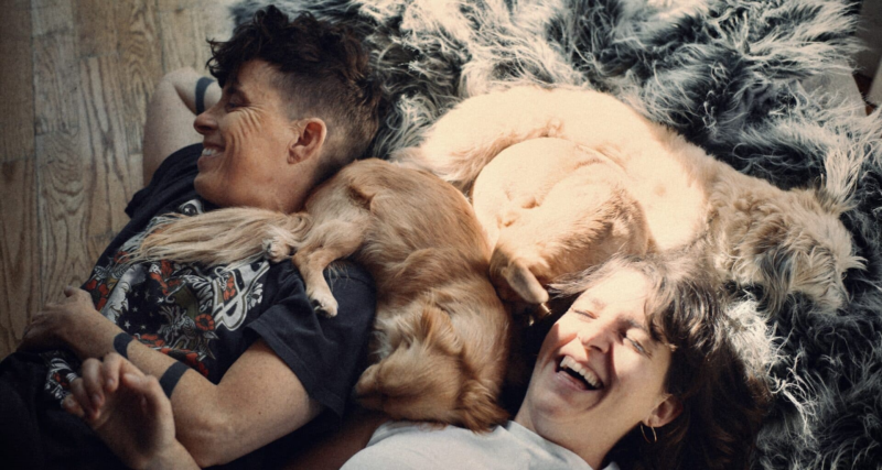 Andrea e Megan estão deitadas em um chão de madeira, com um carpete cinza cheio. Entre elas, estão dois cachorros de pelos loiros dourados. Uma luz bate no pelo de um deles e no rosto de Megan, mulher branca de cabelos castanhos, usando uma blusa branca, que está rindo com os olhos fechados e a cabeça para cima. Andrea, pessoa não binária branca, com cabelos pretos curtos, uma tatuagem no braço e uma camiseta preta estampada, sorri, olhando para o lado.