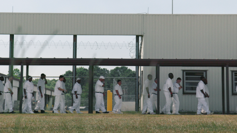 Cena de Alabama: Presos do Sistema. Homens vestidos com uniformes brancos caminham em fila por um corredor ao ar livre cercado por grades e arame farpado, ao lado de um prédio carcerário.