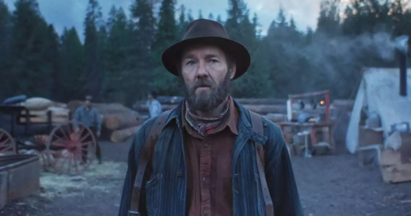 Cena do filme Sonhos de Trem. Na imagem, há um homem branco de cabelos e barba castanho escura. Ele usa três camadas de roupa. Na foto, é possível ver a camisa de botão marrom e uma espécie de jaqueta azul por cima. Atrás dele, há madeiras e árvores. Ele usa um chapéu marrom e está com a feição séria.