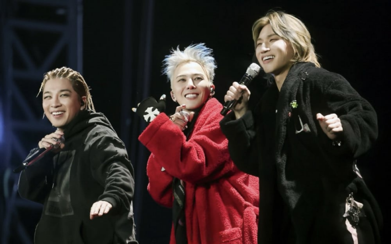 G-DRAGON perforando com Daesung e Taeyang durante sua Turnê Übermensch. Foto retangular com fundo preto. À esquerda temos o cantor Taeyang, um homem asiático com cabelo trançado de cor castanho médio, vestindo um moletom preto. No centro, G-DRAGON, um homem asiático, de cabelo platinado com detalhes em azul, vestindo um roupão vermelho. A direita, Daesung, um homem asiatíco de cabelo castanho claro, liso e médio, vestindo um roupão preto. Os três estão sorrindo. 