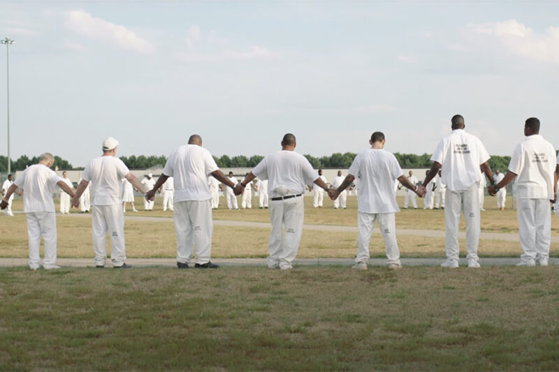 Cena de Alabama: Presos do Sistema. Vários homens usando uniformes brancos estão posicionados em um campo gramado e dão as mãos, formando um grande círculo. No fundo, aparece árvores e o céu azul.
