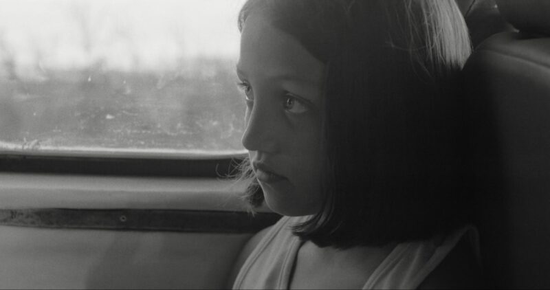 Fotografia em preto e branco. Close do perfil do rosto de uma menina com cabelos lisos e curtos. Ela olha de forma pensativa, com semblante sério, através da janela de um veículo. A luz natural ilumina parte do seu rosto.
