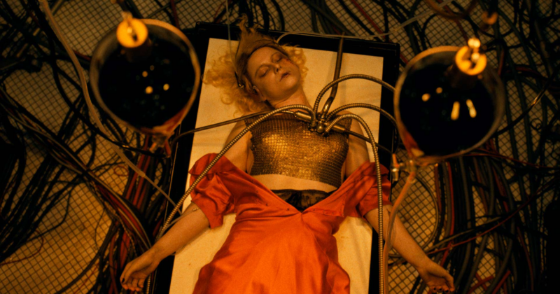 Mulher loira deitada em uma mesa ligada a cabos e equipamentos mecânicos, usando vestido vermelho e peça metálica no peito, em um ambiente industrial com iluminação amarelada. 