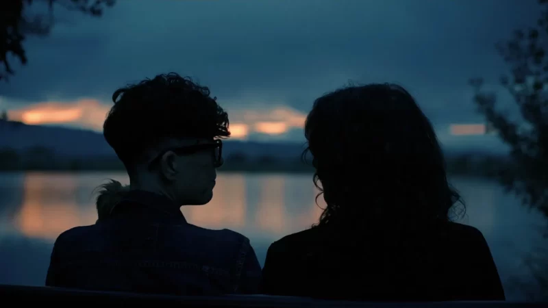 Duas silhuetas escuras aparecem no centro da imagem, são Andrea, pessoa não binária branca, com cabelos pretos curtos, e Megan, uma mulher branca com cabelo castanho médio e franja. Elas olham uma para outra, com os ombros encostados. Ao fundo, vemos uma paisagem em tons azuis de um grande lago e nuvens cobrindo o céu, com algumas árvores aos cantos. Parte do céu aparece rosado como um pôr do Sol, com a luz sendo refletida na água.