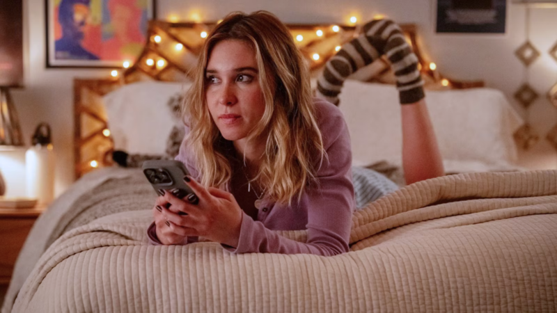 Texto Alternativo: A atriz Isabel May (Tatum Evans) está deitada de bruços em uma cama com roupas de cama claras. Ela veste um casaco lilás e segura um celular com as duas mãos, olhando para o lado com uma expressão de preocupação ou alerta. O quarto está decorado com luzes quentes e pequenos quadros ao fundo.