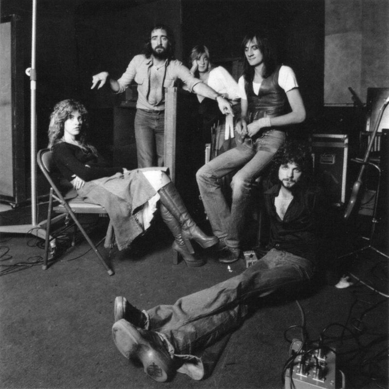 Fotografia em preto-e-branco de cinco pessoas em um estúdio de gravação. À esquerda, uma mulher está sentada em uma cadeira, pernas cruzadas, usando botas de cano alto e uma saia longa. À direita, um homem está sentado no chão, reclinado, com expressão séria. Atrás deles, três pessoas estão de pé: uma mulher ao centro, ligeiramente sorrindo, e dois homens, um com barba e camisa aberta e outro com colete e camiseta branca. Cabos e equipamentos de estúdio aparecem no chão e no fundo.