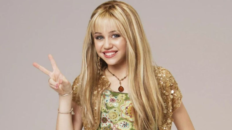 A personagem Hannah Montana, jovem loira sorrindo para a câmera, usando uma blusa com brilho dourado e fazendo sinal de paz com a mão em fundo neutro