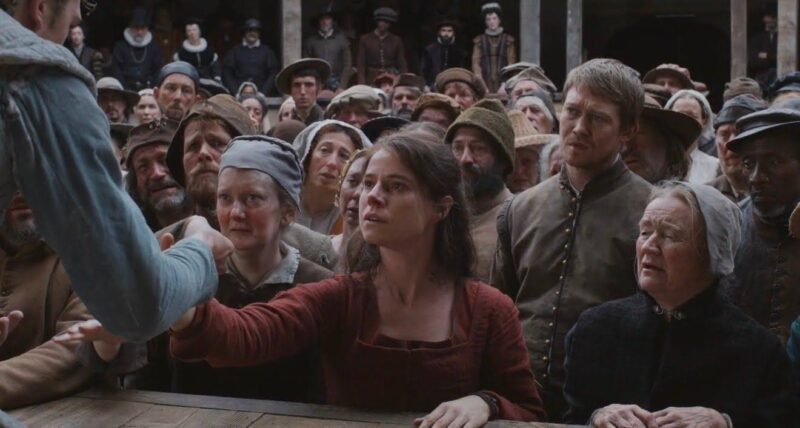 Uma multidão está concentrada diante de uma estrutura de madeira. No centro, uma mulher de cabelos castanhos e vestido vermelho estende a mão em direção a um homem fora de cena. Sua expressão revela concentração e envolvimento emocional. Ao redor, as pessoas observam com atenção. Seus rostos revelam sentimentos variados a respeito do homem. Todos utilizam roupas típicas do século XVI.