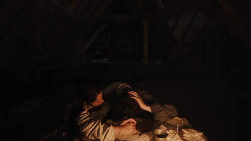 O ambiente é escuro e rústico, iluminado apenas por uma luz quente. Duas pessoas estão sentadas em uma mesa coberta por papéis e um copo. O homem apoia a cabeça na mesa, enquanto a mulher ao seu lado o abraça, com as mãos em seus cabelos. No fundo escuro, há estruturas de madeira e uma cama.