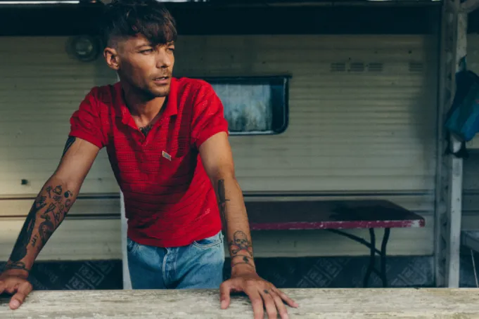  Fotografia de Louis Tomlinson em frente a um trailer, durante o dia. Na foto, ele aparece da cintura para cima, vestindo uma camisa polo vermelha e calça jeans clara. Louis está apoiado com as mãos em uma viga de madeira, olhando para o lado. Seus braços estão estendidos, exibindo várias tatuagens. Ao fundo, vê-se uma mesa vermelha