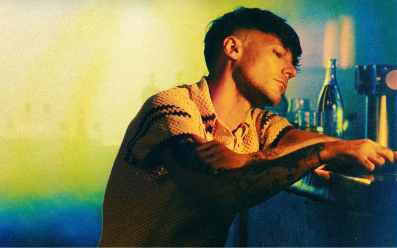  Fotografia de Louis Tomlinson em um ambiente interno com iluminação em tons amarelos e azuis. O cantor, um homem branco de cabelos castanhos curtos e barba rala, aparece de perfil, levemente inclinado em um balcão. Seus braços estão em cima desse balcão e ao seu redor tem uma garrafa e um copo meio cheio. Ele veste uma camisa de tricô bege com listras pretas horizontais e tem diversas tatuagens visíveis nos braços.