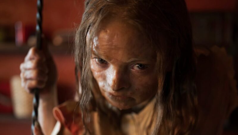 Cena do filme Maldição da Múmia Na imagem, a personagem Katie encara sua frente com um olhar penetrante. Ela é uma garota na faixa dos 17 anos, de pele clara, com textura seca e cicatrizada. Seus cabelos são claros, longos e finos. A mão direita segura uma corda. O fundo é seu quarto, de paredes vermelhas. 