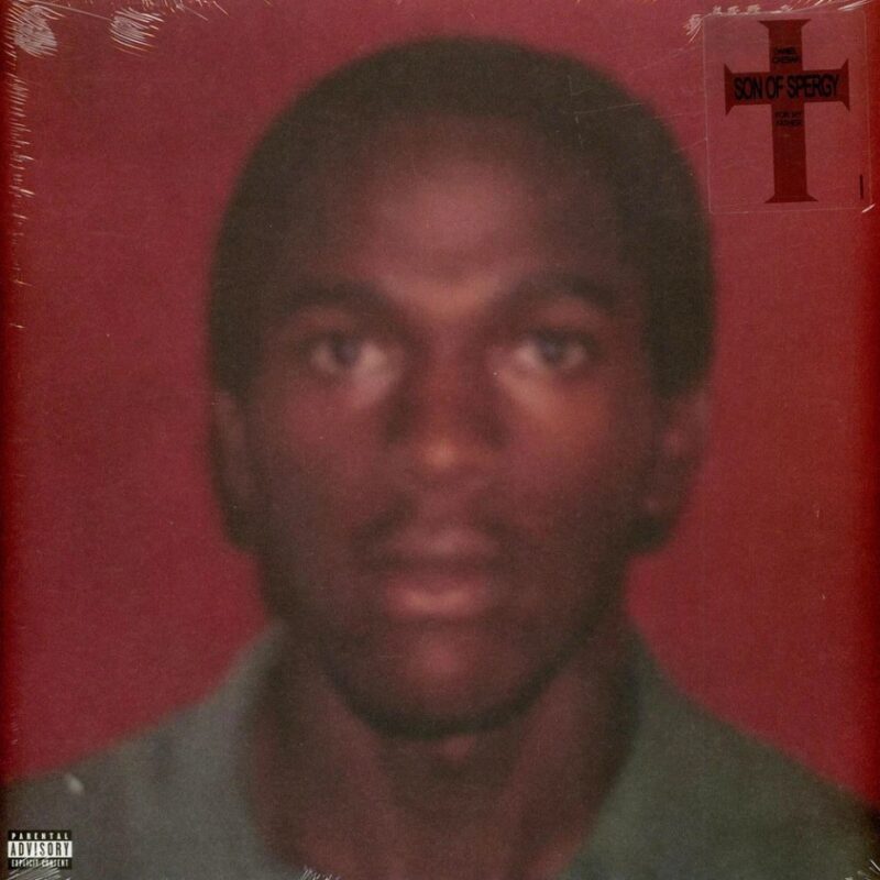 No centro da imagem há um homem de frente para a câmera, com um microfone. Ele está bem distante e nos cantos da imagem há a sombra de pessoas que estão viradas para ele. A foto é tirada dentro de uma catedral durante a noite, com grandes vitrais ao centro sendo iluminados por luzes que vêm do canto inferior direito e do canto inferior esquerdo.