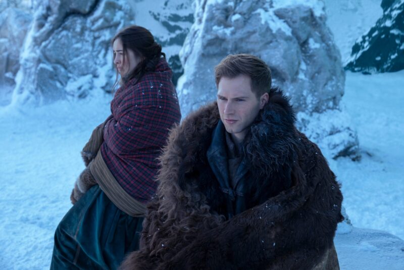 No centro da imagem, um homem e uma mulher estão sentados ao ar livre em um cenário de inverno. Ao redor, há formações rochosas cercadas por neve. O homem, à direita, tem cabelos loiros curtos e veste um manto de pele marrom. Ele olha para frente com uma expressão séria. A mulher, à esquerda, tem cabelos castanhos presos, está em posição de perfil e usa um vestido verde sob um cobertor vermelho. Seu semblante é sério e preocupado.