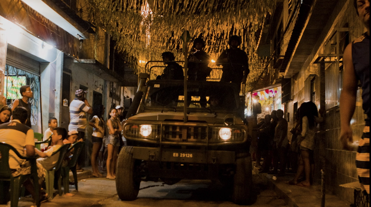 Fotografia noturna de uma rua estreita em uma favela, decorada com bandeirolas que cruzam o alto da via. No centro, um jipe militar escuro com soldados armados avança em direção à câmera. À esquerda, moradores, incluindo crianças e adultos, estão sentados em mesas de plástico ou em pé na calçada, observando a passagem do veículo sob uma iluminação amarelada e densa.
