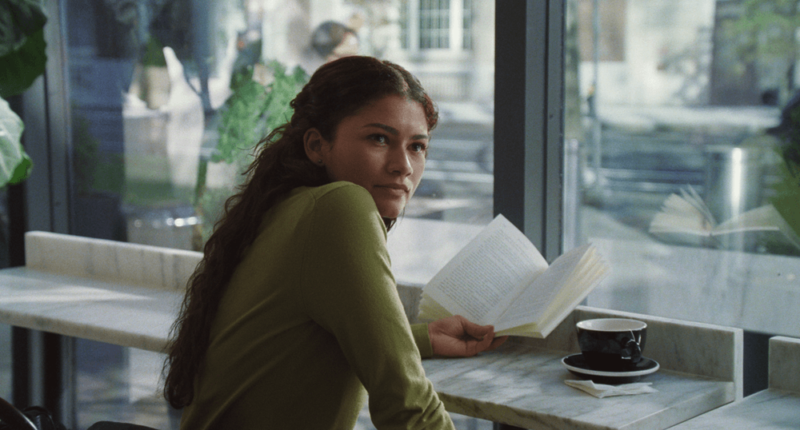 Cena do filme O Drama. Na imagem, há uma mulher negra de cabelos cacheados castanhos. Ela está em uma cafeteria e veste uma blusa verde de manga longa. Ela lê um livro e toma café em uma xícara preta.