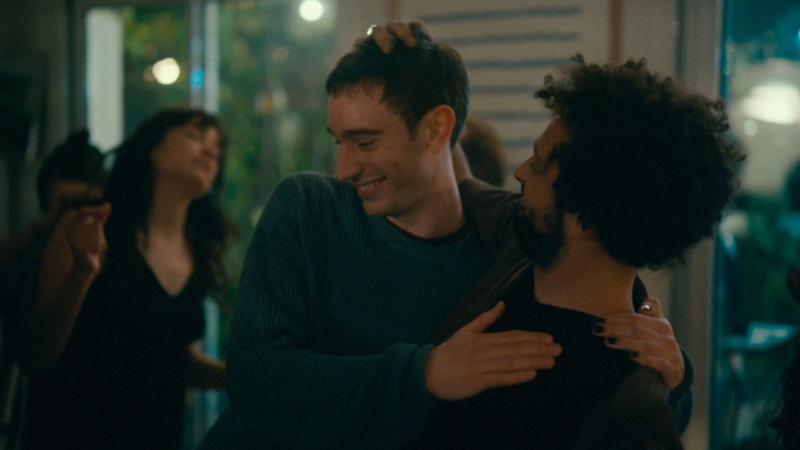 Uma foto cinematográfica colorida e granulada, com profundidade de campo rasa, mostrando o ator Théodore Pellerin como Nino. Ele veste um suéter azul texturizado e sorri suavemente, com a mão na cabeça de outro homem de cabelo afro curto e barba, que está encostado nele em um abraço caloroso. O segundo homem, de jaqueta marrom, está de perfil para a direita. Eles estão em um ambiente de festa interno e difuso, com luzes bokeh quentes ao fundo e outras pessoas dançando, desfocadas e de costas para a câmera, criando uma atmosfera terna e íntima.