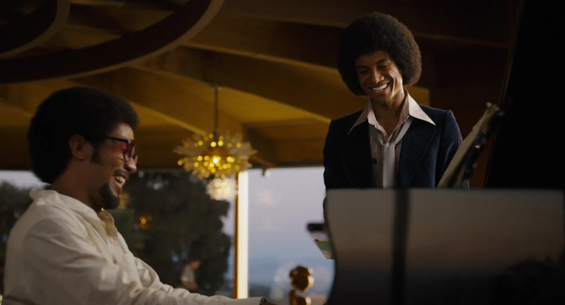 Cena do filme MichaelÀ direita, Jaafar, como Michael, sorri amplamente enquanto está em pé ao lado de um piano de cauda. Ele usa um terno escuro e camisa de gola larga. À esquerda, Kendrick Sampson, como Quincy Jones, sentado ao piano sorri de volta para ele. O ambiente tem uma arquitetura contemporânea dos anos 1970, com uma luminária dourada pendurada ao fundo. 