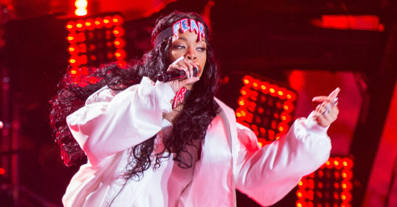 Fotografia de plano médio da cantora Rihanna durante uma performance ao vivo. Ela segura um microfone com a mão direita e gesticula com a esquerda. Rihanna veste uma jaqueta puffer branca volumosa e uma faixa preta na cabeça com a palavra "FEAR" escrita em letras azuis e vermelhas com efeito de "escorrido". O fundo é composto por luzes de palco vermelhas intensas e desfocadas, criando uma atmosfera quente e energética. O cabelo dela está solto, longo e cacheado.