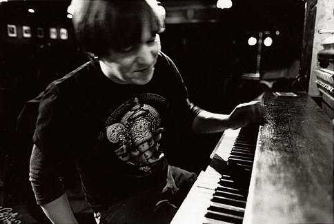 Fotografia retangular horizontal em preto e branco. A imagem foca em um piano ao lado direito no primeiro plano. Elliott Smith está sentado diante dele, posicionado na lateral esquerda da imagem. Ele é um homem branco, magro, com cabelo castanho curto, inclinado em direção às teclas. Sua mão direita está suspensa próxima ao teclado, enquanto seu corpo se inclina para frente. Ele veste uma camiseta escura com estampa desenhada no centro. O fundo é escuro e desfocado, sugere um ambiente de estúdio musical.