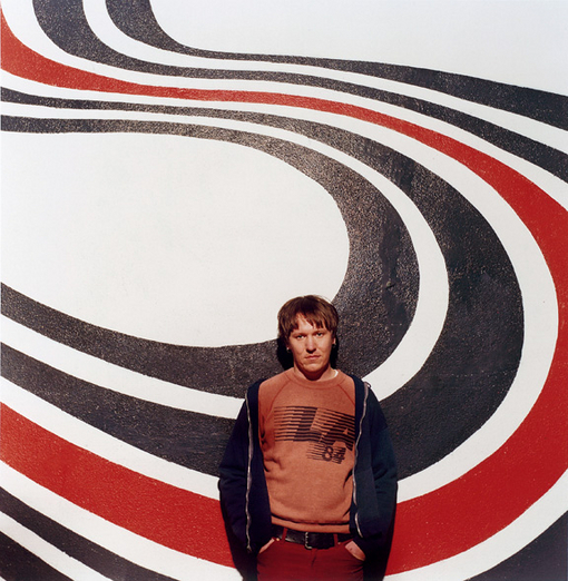 Fotografia quadrada colorida. Elliott Smith está em pé, no centro da imagem, de frente para a câmera. Um homem branco, com cabelo castanho curto e expressão neutra. Veste camiseta marrom com estampa no peito, jaqueta escura aberta e calça vermelha. Atrás dele há um grande mural pintado com faixas curvas seguindo um padrão nas cores preta, branca e vermelha, em alto contraste, que ocupam todo o fundo da imagem. O ambiente é externo.