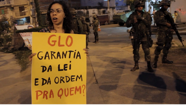 Uma mulher de cabelos cacheados e óculos segura um grande cartaz amarelo em primeiro plano. No cartaz, está escrito em letras garrafais: "GLO GARANTIA DA LEI E DA ORDEM PRA QUEM?". Ao fundo, em uma área urbana aberta durante a noite, soldados fardados portando fuzis e um veículo blindado (tanque) compõem o cenário de vigilância.