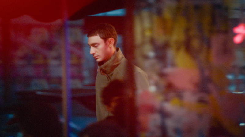 Uma foto cinematográfica granulada de ângulo fechado do ator Théodore Pellerin no papel de Nino. Ele veste uma jaqueta verde oliva e está de perfil para a esquerda, olhando para baixo com uma expressão pensativa e contida. O primeiro plano e o fundo estão borrados por luzes de neon vermelhas e azuis e silhuetas desfocadas, emoldurando o rosto de Nino e criando uma sensação de isolamento e introspecção em um ambiente urbano noturno.