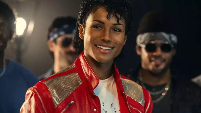 Cena do filme MichaelJaafar Jackson, como Michael Jackson, sorrindo e olhando em direção ao lado esquerdo da foto. Ele usa a famosa jaqueta de couro vermelha com detalhes prateados nos ombros e uma camiseta branca com estampa de teclado de piano. Ao fundo, figurantes e luzes de estúdio estão desfocados, sugerindo o set de gravação de um videoclipe. 
