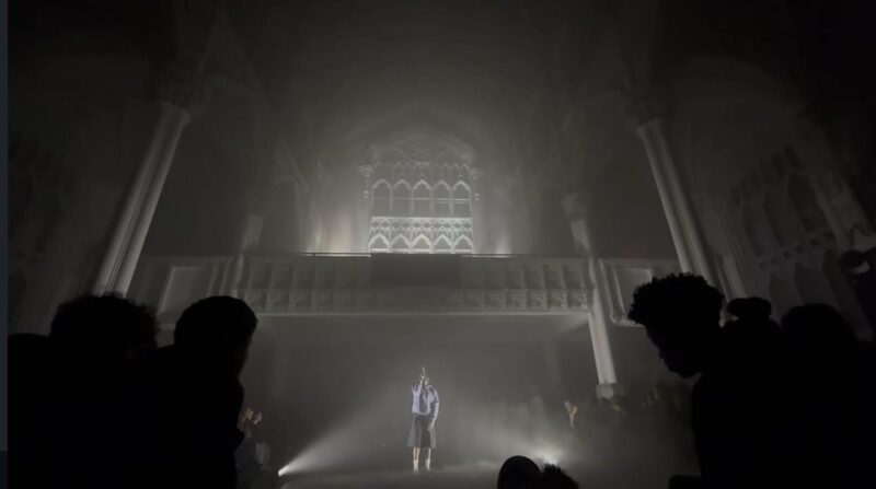A imagem apresenta um retrato de um homem jovem fotografado contra um fundo vermelho liso e intenso. Ele está centralizado na foto, olhando diretamente para a câmera, com expressão neutra e séria. O homem tem cabelo curto, e veste uma camisa de colarinho verde escuro, ligeiramente aberta no topo. A imagem possui uma aparência antiga. O enquadramento é simples, mostrando apenas a cabeça e parte dos ombros do retratado.