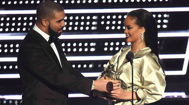 Fotografia de perfil de Drake e Rihanna no palco do VMA. À esquerda, Drake, um homem negro com barba curta e cabelo raspado, veste um smoking preto clássico com gravata borboleta e olha para Rihanna com um sorriso leve. À direita, Rihanna, uma mulher negra com cabelos longos e lisos penteados para trás, usa um vestido longo de cetim dourado pálido e brincos de diamante pendentes. Ambos seguram juntos um troféu. O fundo é composto por linhas horizontais de luzes brancas brilhantes sobre um painel escuro.