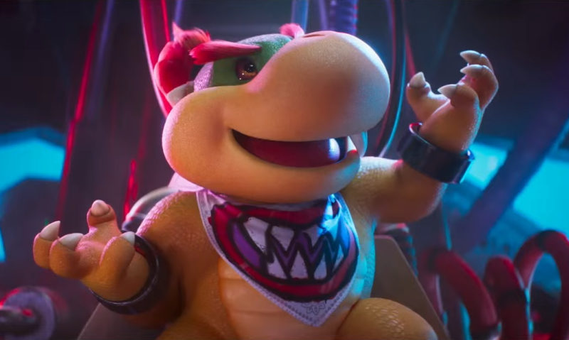 Criatura animada semelhante a um filhote de tartaruga com corpo robusto, pele amarelada e focinho arredondado aparece em primeiro plano. Ela usa uma bandana com desenho de dentes afiados e pulseiras escuras nos braços. O personagem está sentado em uma cadeira metálica com cabos ao redor, levantando as mãos com expressão animada. A iluminação mistura tons de vermelho e azul, criando um ambiente tecnológico e levemente ameaçador.