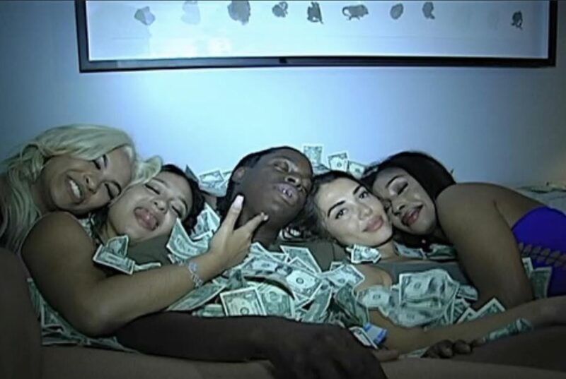 Na imagem, cinco pessoas estão deitadas lado a lado em um sofá, em um ambiente interno com iluminação azulada e atmosfera de festa. Da esquerda para a direita, há quatro mulheres, uma loira e três morenas, e um homem ao centro. As mulheres exibem expressões alegres e descontraídas, algumas sorriem amplamente, enquanto a segunda mostra a língua e faz um gesto de arma com os dedos. O homem, no meio do grupo, tem os olhos fechados e parece relaxado, com notas de dinheiro sobre o peito e o rosto.