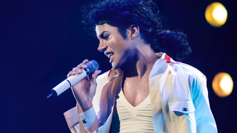 Cena do filme MichaelFotografia de plano médio de Jaafar Jackson como Michael Jackson em um palco. Ele está de perfil, com o corpo levemente inclinado, cantando em um microfone prateado. Ele veste uma camisa branca aberta sobre uma camiseta branca, pele suada sob luzes de palco azuis e douradas. Seu cabelo é preto, cacheado e está preso em um rabo de cavalo baixo. 