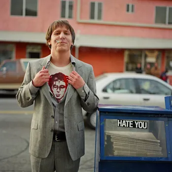 Fotografia quadrada colorida. Elliott Smith está posicionado em uma calçada ao lado esquerdo da imagem, de frente para a câmera. Ele é um homem branco, com cabelo castanho curto e expressão alegre, com um sorriso sutil no rosto. Veste um terno cinza claro, uma camisa também cinza claro e uma camiseta branca com estampa vermelha, as mãos seguram as lapelas do paletó, abrindo-o. O homem está com o cotovelo apoiado em uma caixa de jornal azul com a frase “hate you” escrita em letras brancas, posicionada ao lado direito da imagem. Ao fundo, aparecem carros estacionados e um prédio de fachada rosada, indicando um ambiente urbano. A luz é natural e as cores são suaves.