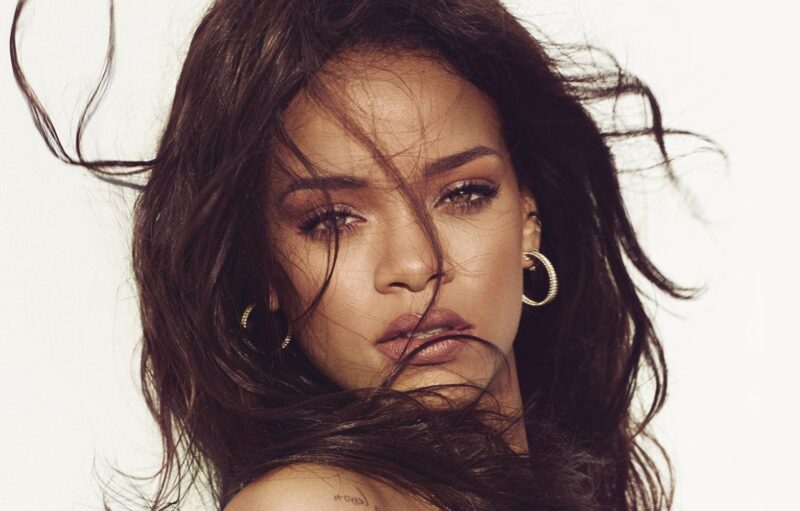 Retrato em primeiríssimo plano (close-up) da cantora Rihanna contra um fundo branco infinito. Ela olha diretamente para a câmera com uma expressão serena e lábios levemente entreabertos. Seu cabelo castanho escuro está solto e bagunçado, com várias mechas sopradas pelo vento cruzando seu rosto. Ela usa brincos de argola dourados com textura trançada e uma maquiagem em tons terrosos e naturais. Parte de uma tatuagem é visível em seu ombro esquerdo.