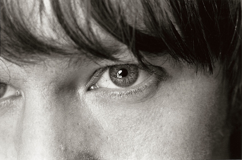 Fotografia retangular horizontal em preto e branco. A imagem mostra um close no rosto de Elliott Smith, enquadrado do nariz para cima. O foco está em seu olho direito, aberto e voltado levemente para a esquerda da imagem, com expressão concentrada. Fios da franja de cor escura caem sobre a testa. A textura da pele é visível, com poros, linhas abaixo dos olhos e pequenas marcas em destaque. A luz é suave, criando contraste delicado entre sombras e áreas claras.