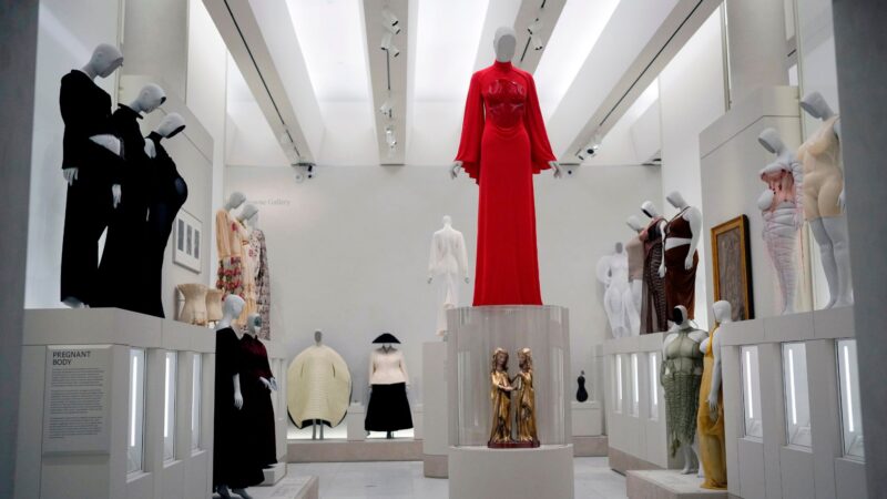 Em uma galeria ampla e iluminada, diversos manequins exibem peças de roupas em tons variados. No centro do espaço, um vestido longo vermelho se destaca sobre uma plataforma elevada, cercado por roupas esculturais e silhuetas experimentais distribuídas pelas paredes brancas do ambiente. 